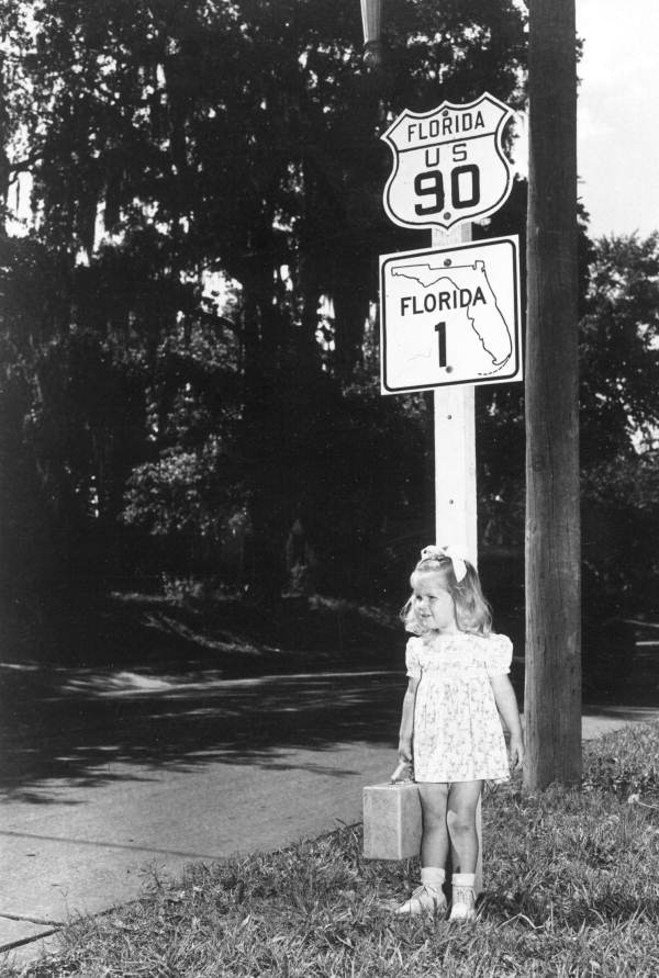 A young girl stands in front of one of the old-style U.S. highway shields for U.S. 90 (1946).