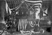 Group portrait of Governor Napoleon P. Broward and ex-Governor Francis P. Fleming standing with other men and Civil War battle flags at the Capitol House chamber.