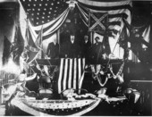 Group portrait of Governor Napoleon P. Broward and ex-Governor Francis P. Fleming standing with other men and Civil War battle flags at the Capitol House chamber.