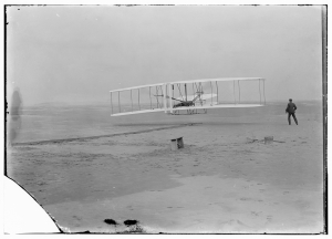 First powered flight in a large field with Orville Wright lying flat on the lower wing of a biplane while Wilbur Wright runs alongside the plane.