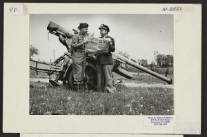 Two men standing by a large German anti-aircraft gun at Verdun, France, reading a newspaper with the headline Victory.