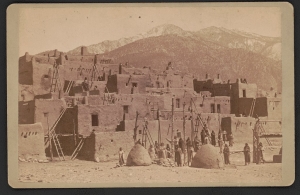 Multistoried adobe pueblo with a group of Native people posing in the foreground.