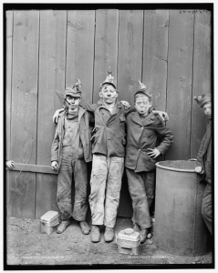 Three coal breaker boys in worn clothing with soot on their faces embracing in front of a wood wall.