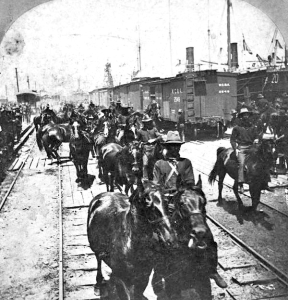 The Rough Riders, or the 1st United States Volunteer Cavalry, arrive in Tampa with a view of the docks as men on horseback ride toward the camera.