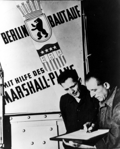 Two workmen looking over a chart with a large sign behind them with German writing.