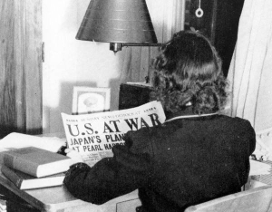Over-the-shoulder view of a woman reading a newspaper with the visible headline U.S. At War alongside a partially visible subtitle about Japan's planes attacking Pearl Harbor.
