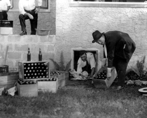 Men in suits removing crates of alcohol from a small basement window with cases stacked on the ground.
