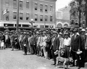 A crowd of men in suits lined up in the street as several onlookers watch out of the windows of buildings in the background.